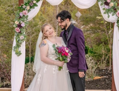 Bride and groom standing together under a floral wedding arch, with the bride holding a bouquet and the couple sharing a quiet moment during an outdoor ceremony.