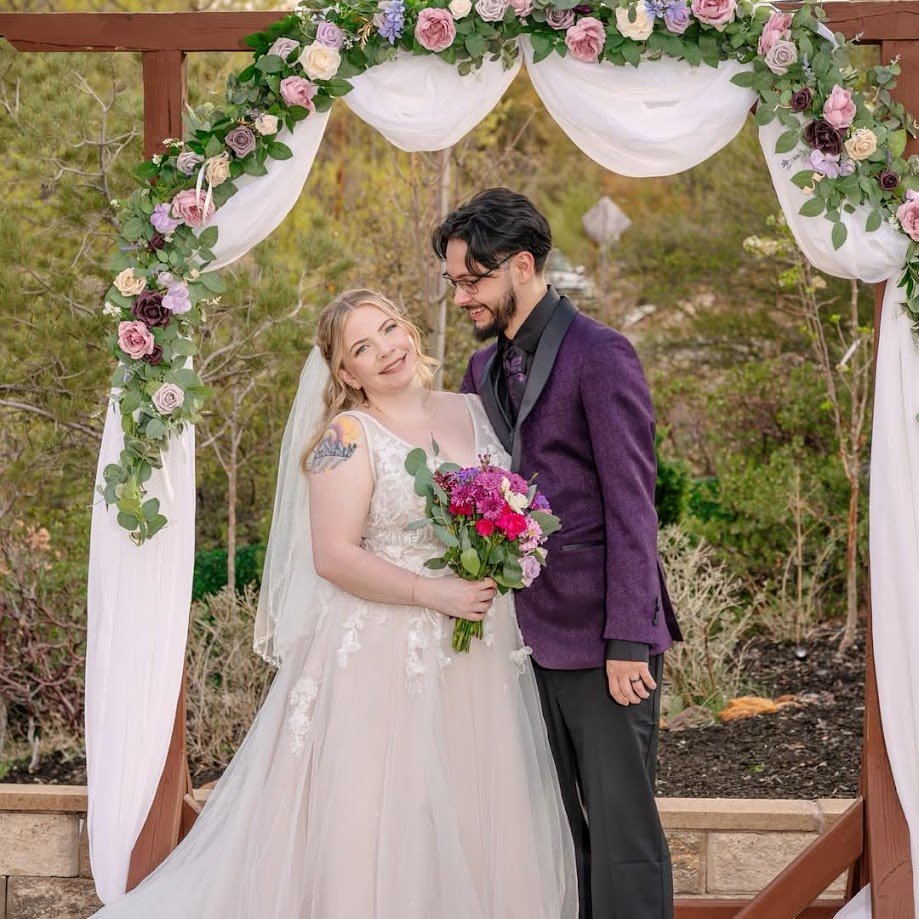 Bride and groom standing together under a floral wedding arch, with the bride holding a bouquet and the couple sharing a quiet moment during an outdoor ceremony.