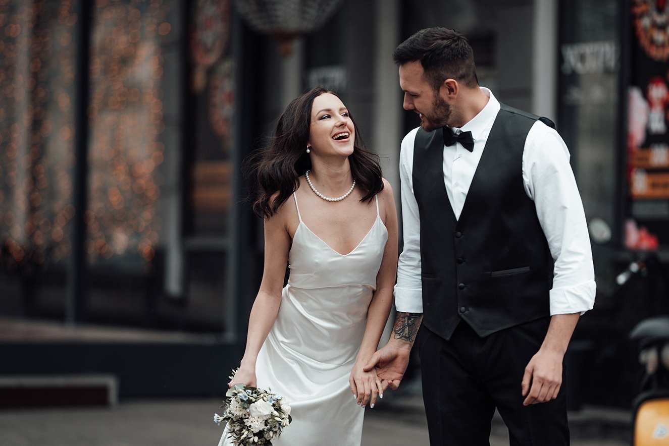 A bride in a white dress and a groom in a black vest and bow tie walk hand in hand down a city street, smiling at each other.